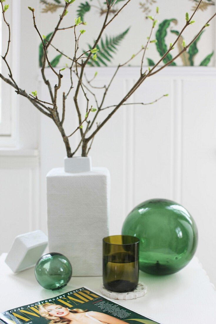 Branches of leaves in white ceramic vase and green drinking glass between two green glass spheres on white table