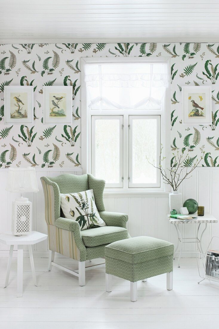 Reading chair with matching footstool below window; white wainscoting below decorative wallpaper with pattern of fern