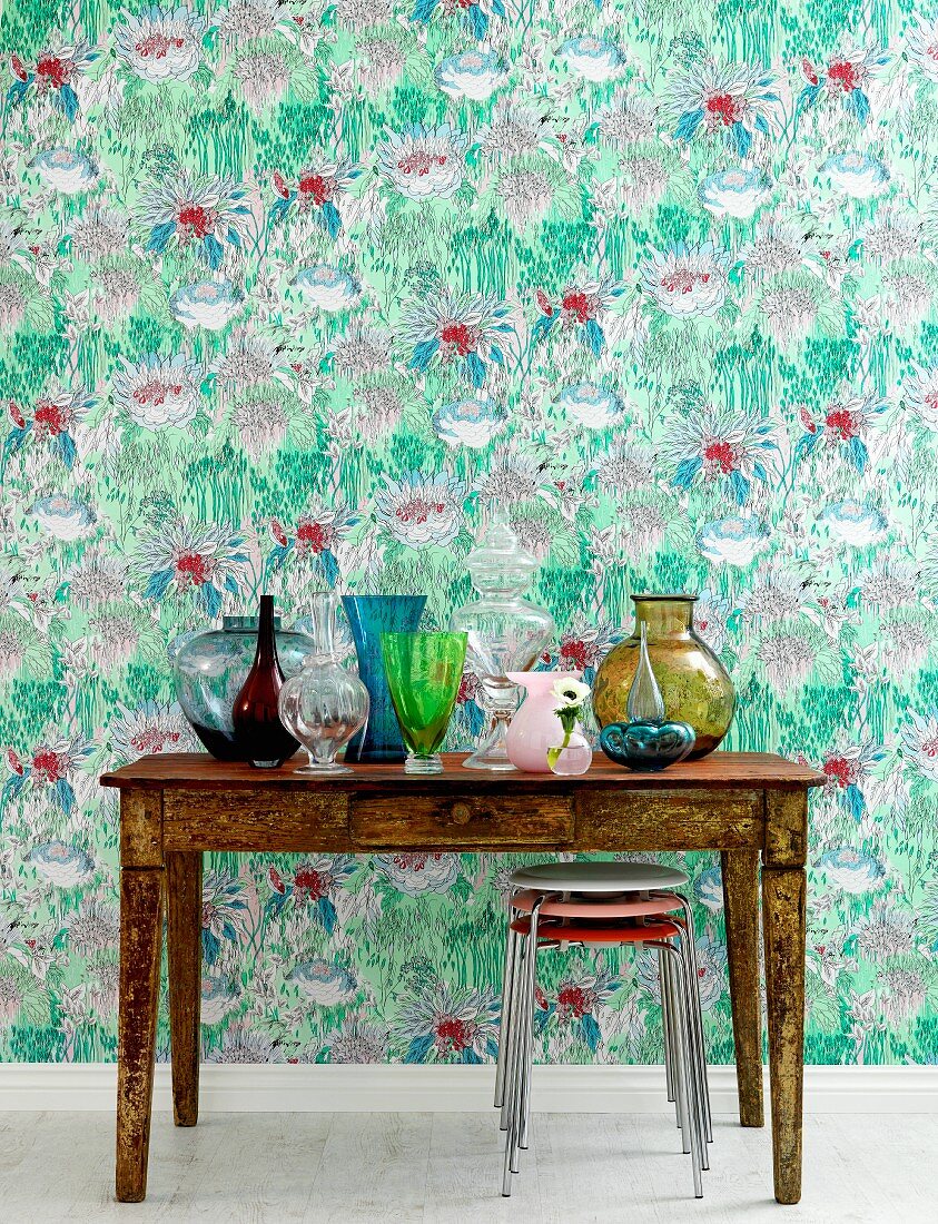 Holztisch in Vintage Stil mit bunter Glasvasen, vor Wand mit malerischem Blumenmuster auf Tapete