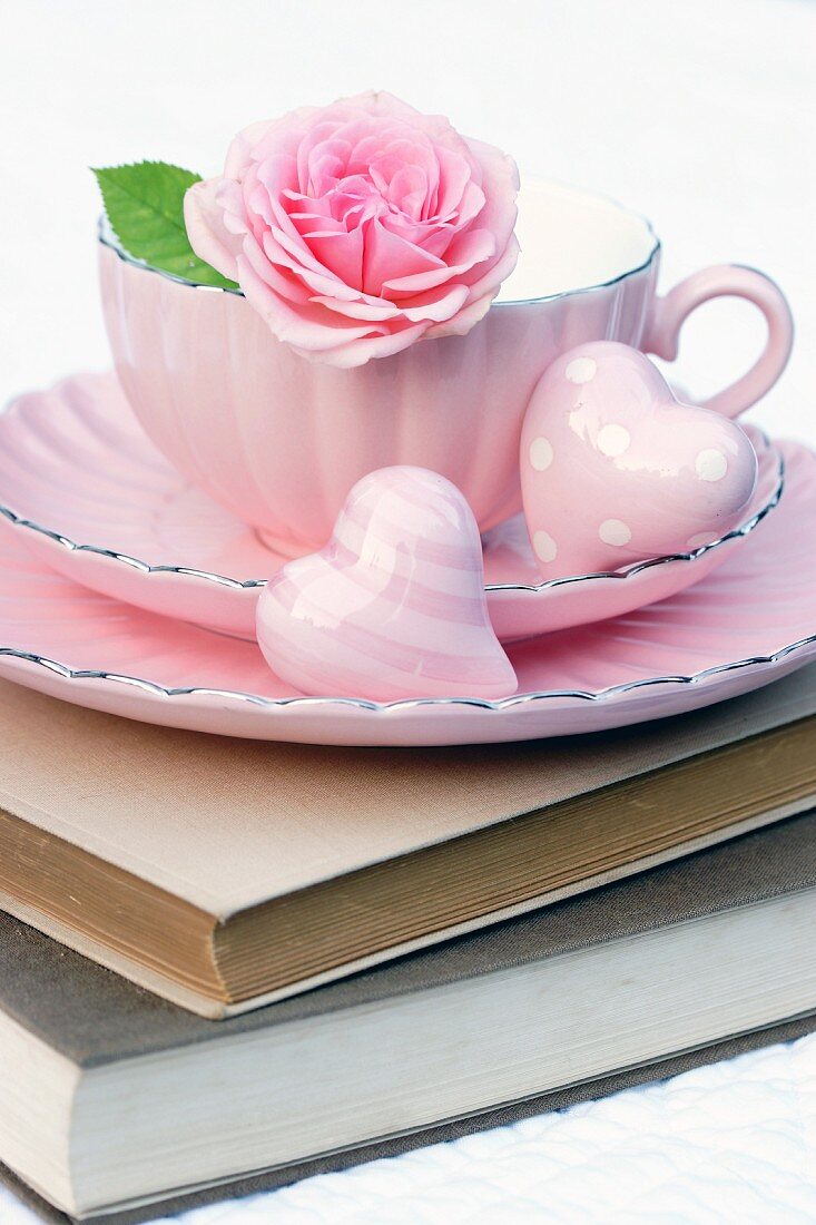 Rosafarbene Tasse auf Bücherstapel dekoriert mit Rosenblüte & Dekoherzen