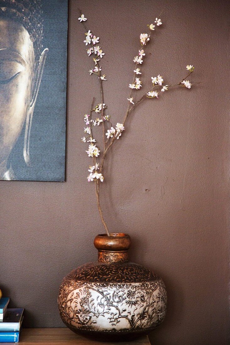 Verzierte Holzvase mit weißem Blütenzweig und teilweise sichtbares Bild vor brauner Wand