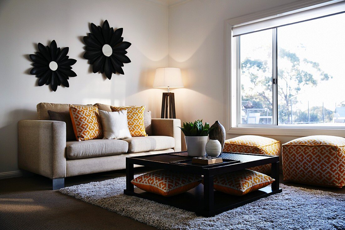 Quadratischer Couchtisch vor Sofa mit gemusterten Kissen und Polsterhockern mit gleichem Muster in Zimmerecke vor Fenster, an gegenüberliegender Wand Spiegel mit blumenförmigem Rahmen