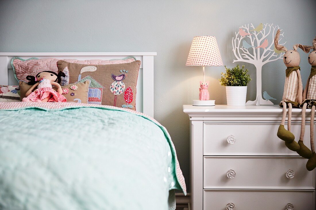 Mädchenzimmer - Stoffpuppe auf besticktem Kissen im Bett mit Kopfteil, neben weisser Kommode mit Tischleuchte und Stofftieren