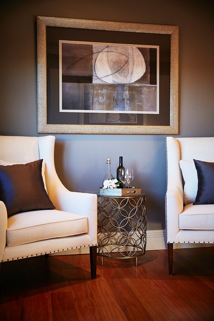 Filigraner Beistelltisch aus Metallgitter zwischen eleganten, weissbezogenen Sesseln vor hellgrauer Wand mit gerahmtem Bild