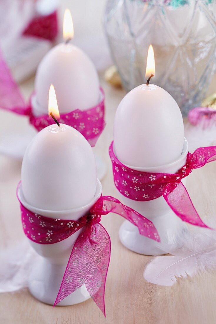 Egg-shaped candles in white egg cups decorated with pink ribbons