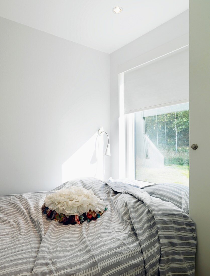 Window with roller blind at foot of bed, floral, ruched scatter cushions on striped grey cover