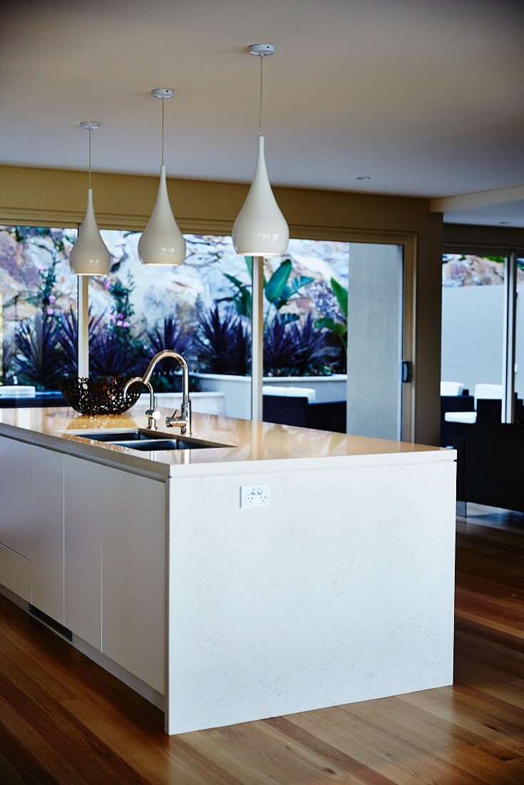 weiße Kücheninsel mit eingebauter Spüle unter Leuchtengruppe mit glänzendem, flaschenförmigem Schirm in modernem Wohnraum mit raumhohen Terrassentüren