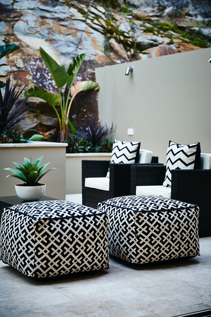 Gemusterte Polsterhocker und schwarze Sessel mit gemusterten Kissen auf Terrasse, neben Hochbeeten mit tropischen Pflanzen teilweise vor Felswand und gemauerter Wand