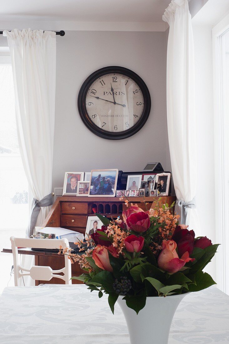 Blumenstrauss in Porzellanvase auf Tisch, im Hintergrund antike Wanduhr über Sekretär