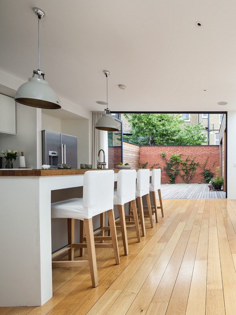 Barhocker mit weissen Hussen vor Küchenblock in offener Designerküche, im Hintergrund geöffnete Terrassen Falttür