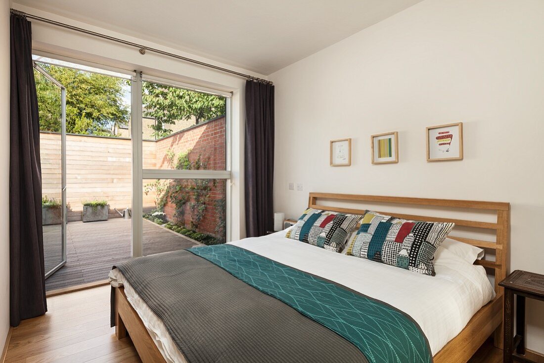 Doppelbett mit Holzrahmen in schlichtem Schlafzimmer, seitlich Fensterfront mit offener Terrassentür