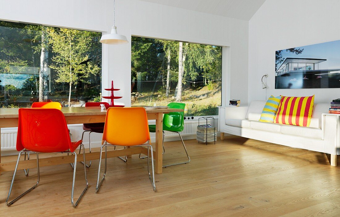 Bunte Schalenstühle und Holztisch vor Panoramafenster mit Waldblick; seitlich Sofa mit bunten Streifenkissen