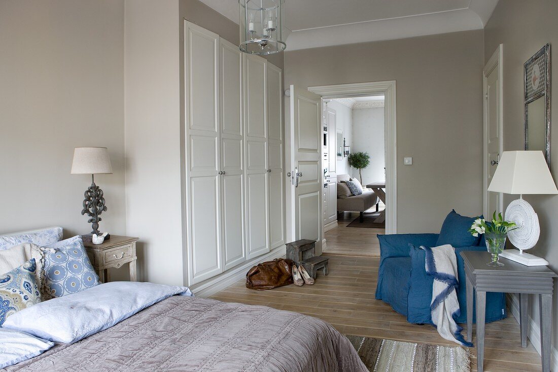 Klassisches weißes Schlafzimmer mit Naturtönen und blauen Akzenten; Blick vom Bett auf Einbauschrank und Essplatz im Nebenzimmer
