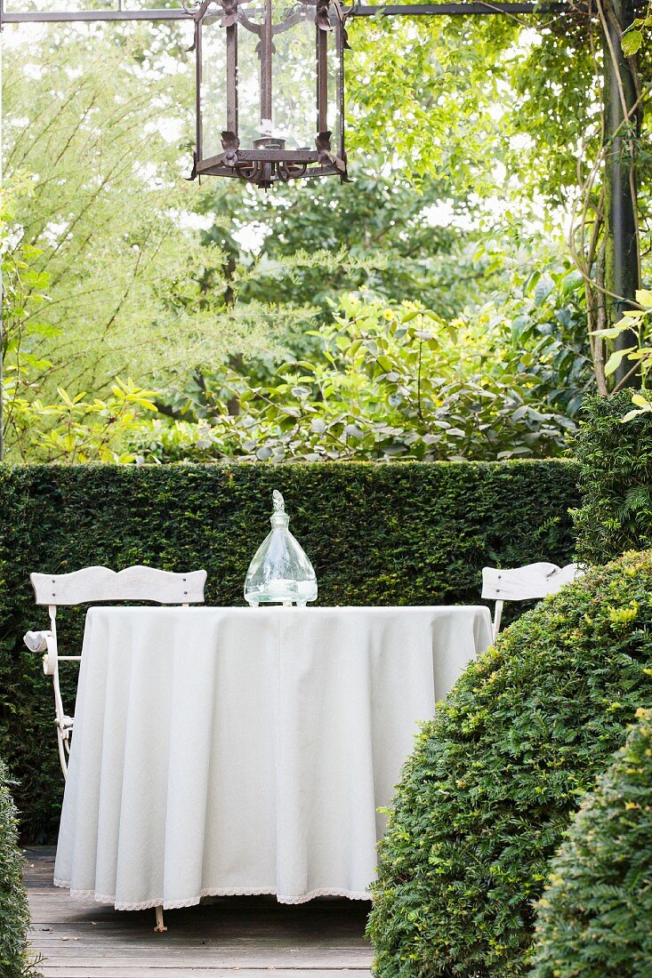Table with long, white tablecloth and garden chairs in front of half-height hedge