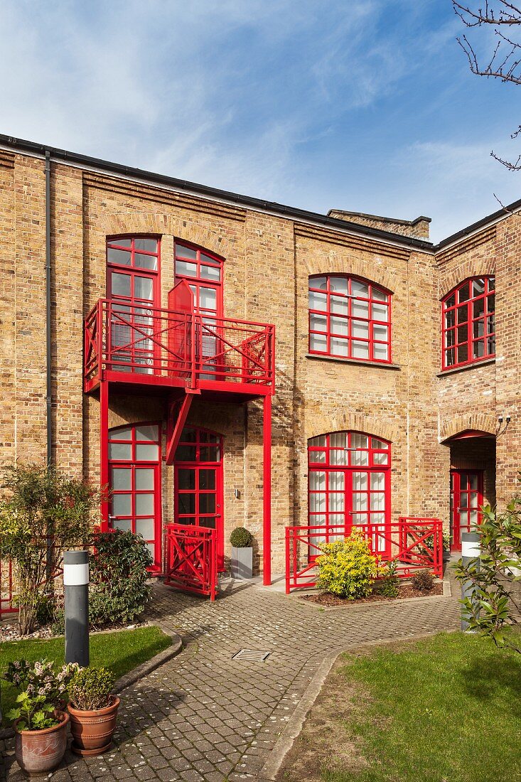 Ehemalige Fabrik als Wohnhaus, rot gestrichene Fensterrahmen und Balkongeländer, davor Innenhof mit eingefassten Beeten und Pflasterwegen