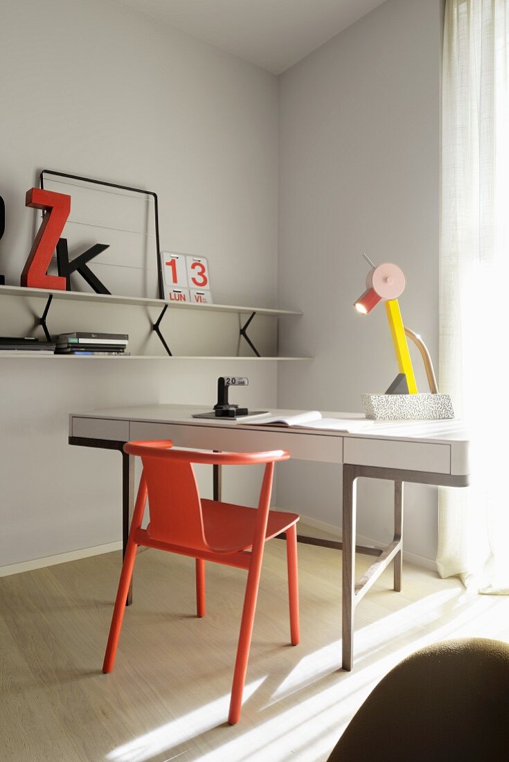 Red classic wooden chair at white writing desk with postmodern table lamp; white shelf on wall painted pale grey