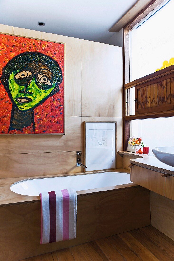 Elegantes Bad mit massgefertigten Holzeinbauten - Badewanne vor Holzpaneelwand mit modernem Bild neben Waschtisch mit Schubladen vor Fenster