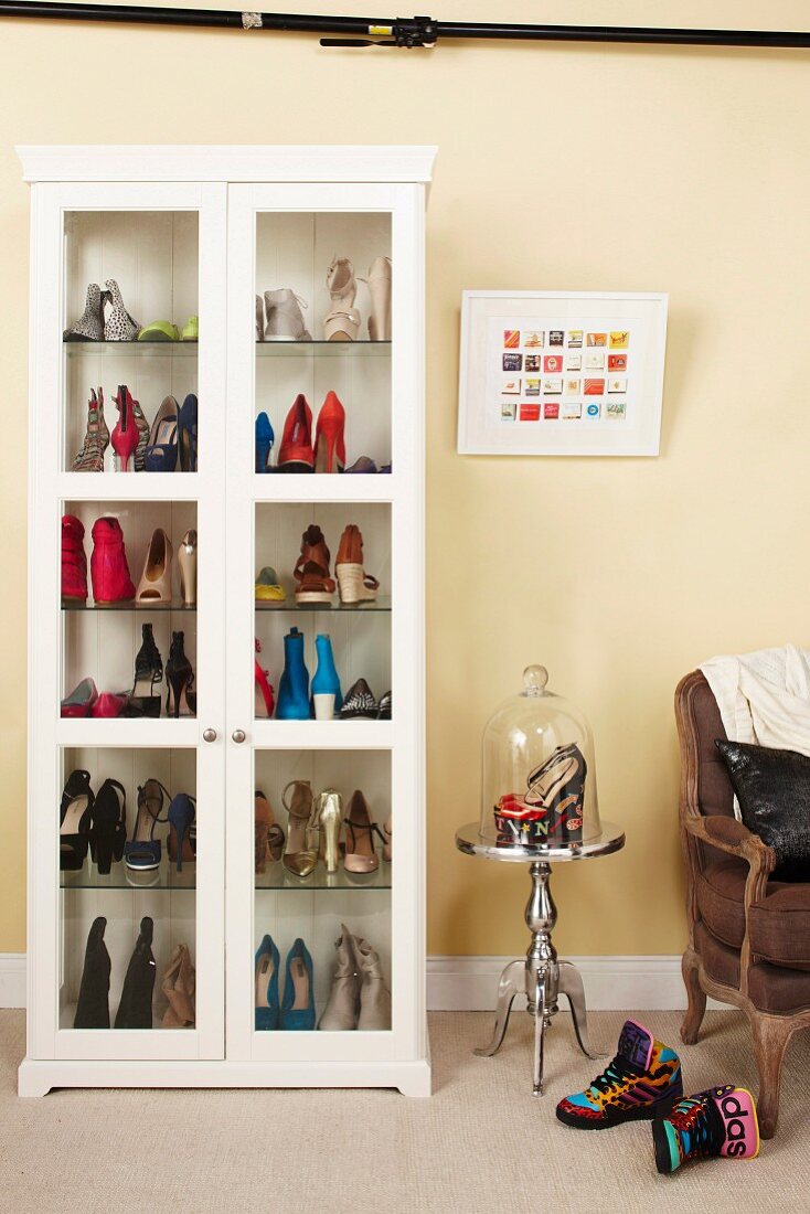Ladies' shoes in white display case next to high heels under glass cover on side table and armchair against wall painted pale yellow