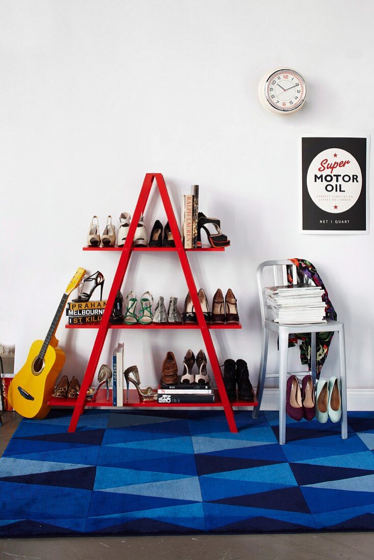 Gitarre an leiterförmiges, rotes Regal mit Damenschuhen und Metall Barhocker unter Poster an weisser Wand, auf Boden Teppich mit geometrischem Muster in verschiedenen Blautönen