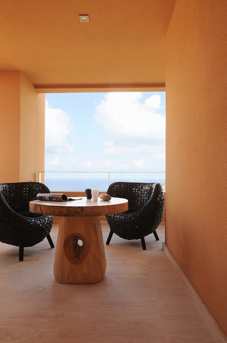 Black wicker chairs and mushroom-shaped, solid wooden table on roofed terrace; sea view through glass balustrade