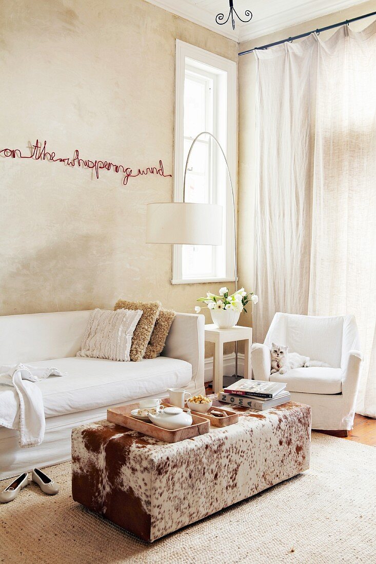 Text artwork on sand-coloured wall above sofa with white loose cover and ottoman with animal-skin cover
