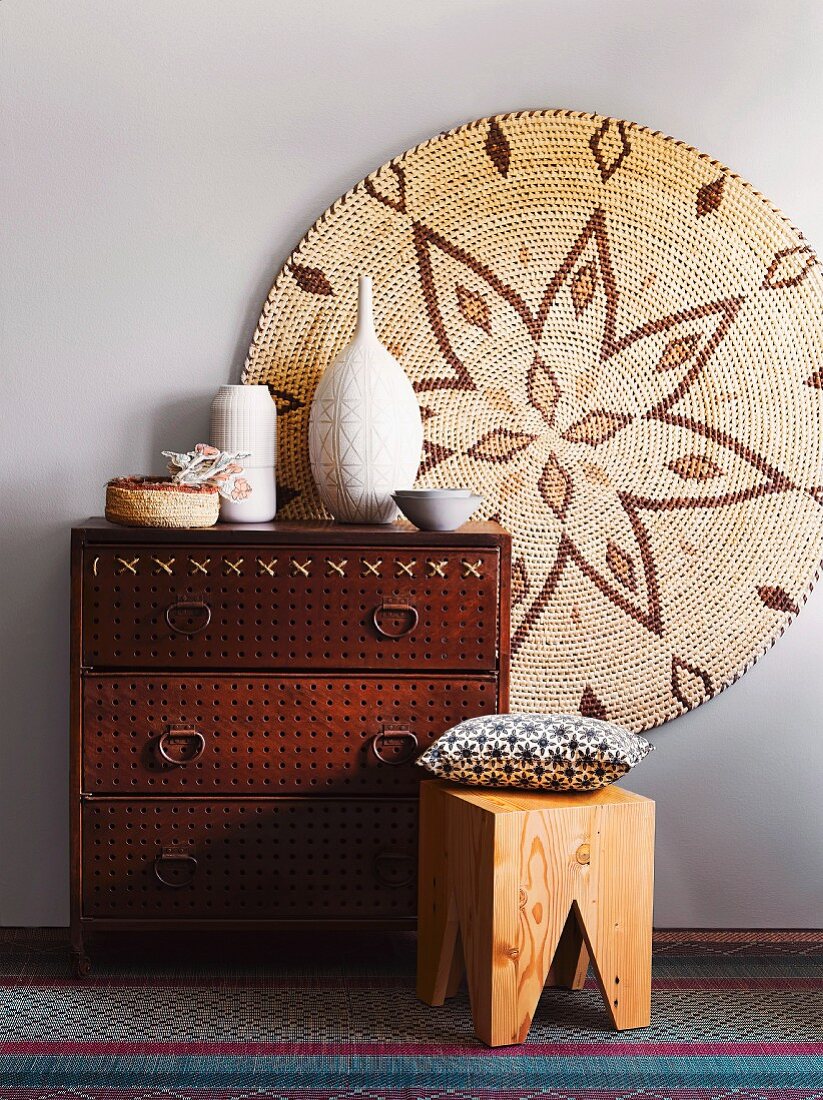 Ethnic arrangement in natural shades - chest of drawers with perforated front, wooden stool and painted picture on wicker disc