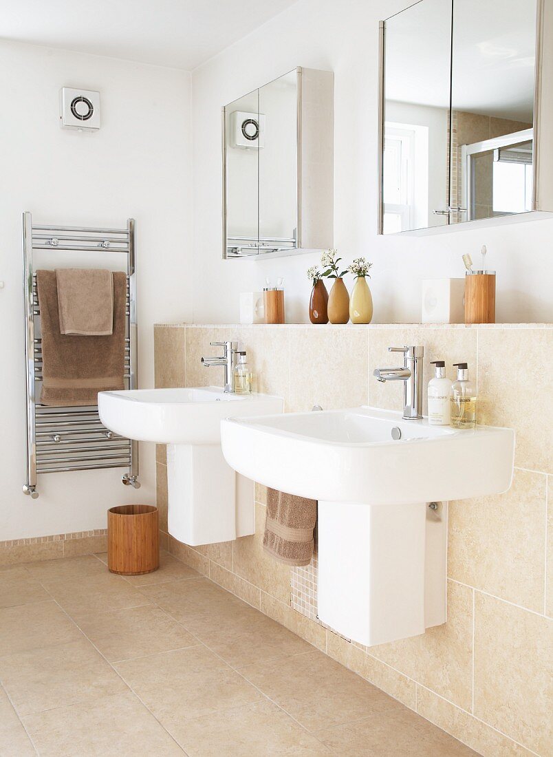 Zwei Einzel Waschbecken an Vormauerung mit sandfarbenen Fliesen, darüber Spiegelschränke an Wand, in der Ecke Edelstahl Handtuchtrockner mit braunen Handtüchern