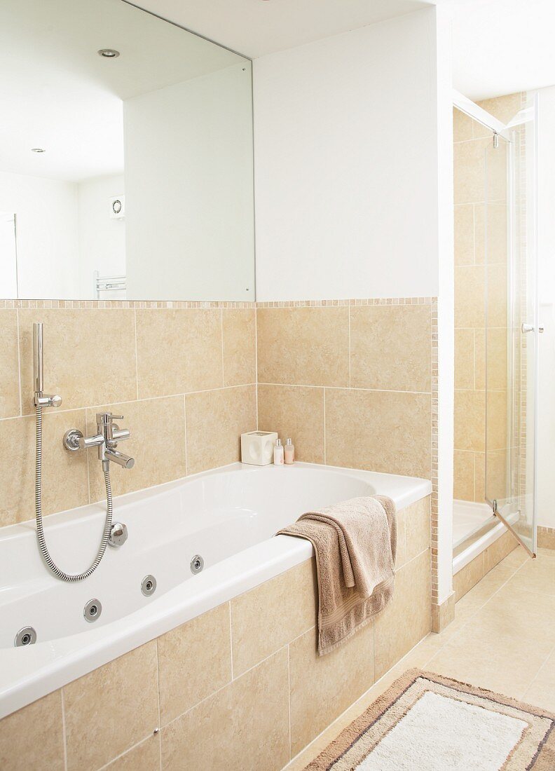 Eingebaute Badewanne vor halbhoher, sandfarbener Fliesenwand, darüber vollflächiger Spiegel an Wand, abgemauerte Dusche mit Glastür im Hintergrund