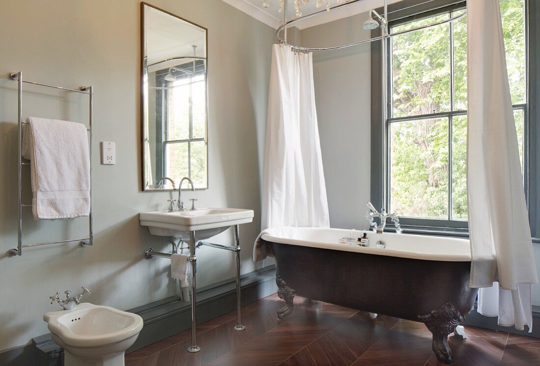 Freistehende Vintage Badewanne mit Duschvorhang vor Fenster, daneben Waschbecken auf Metallgestell unter Wandspiegel im Bad
