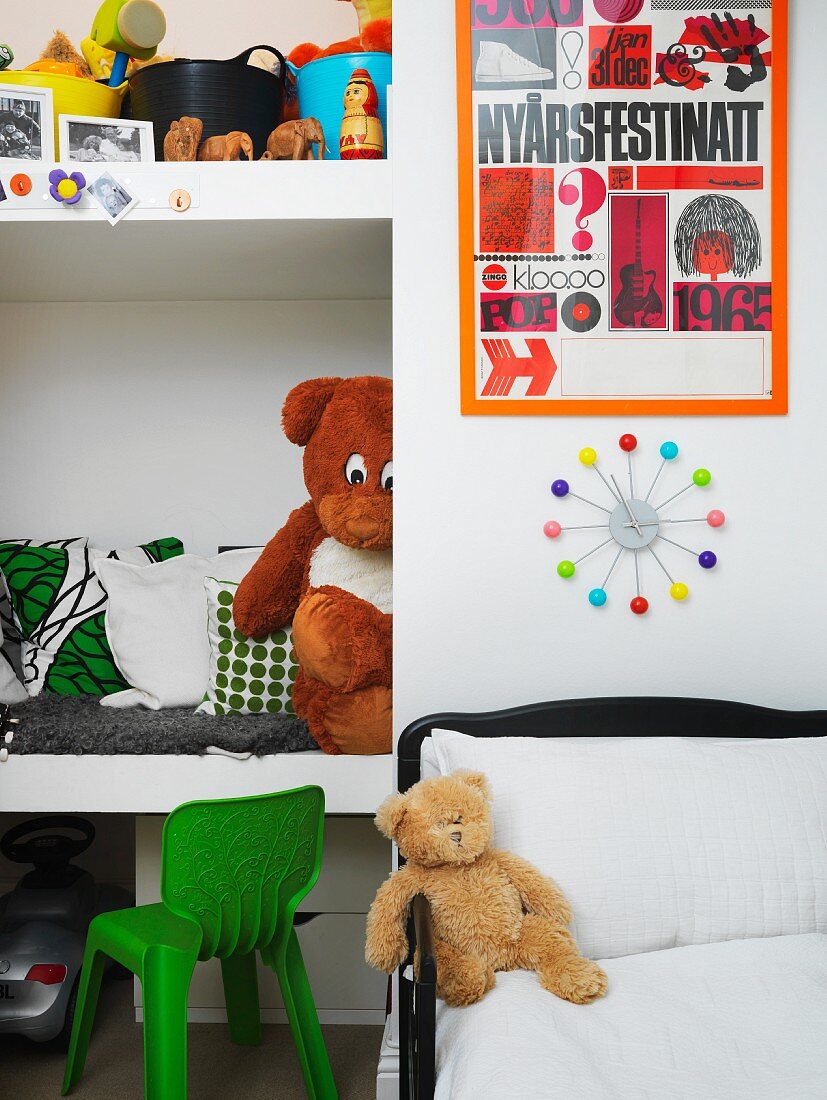 Child's bed below clock on wall next to fitted shelves of cushions and toys in niche