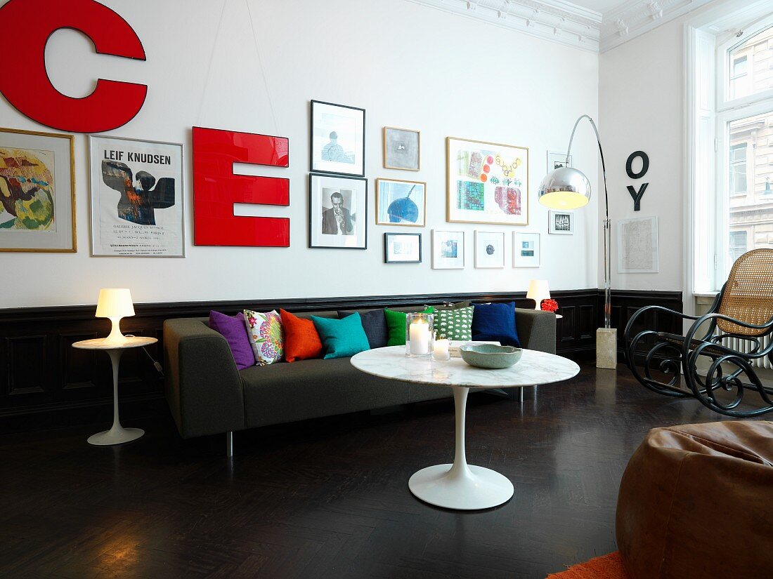 Tulip Tisch, grauer Couch mit bunten Kissen und Thonet Schaukelstuhl in Wohnzimmer mit Bildergalerie und Deko-Buchstaben an der Wand