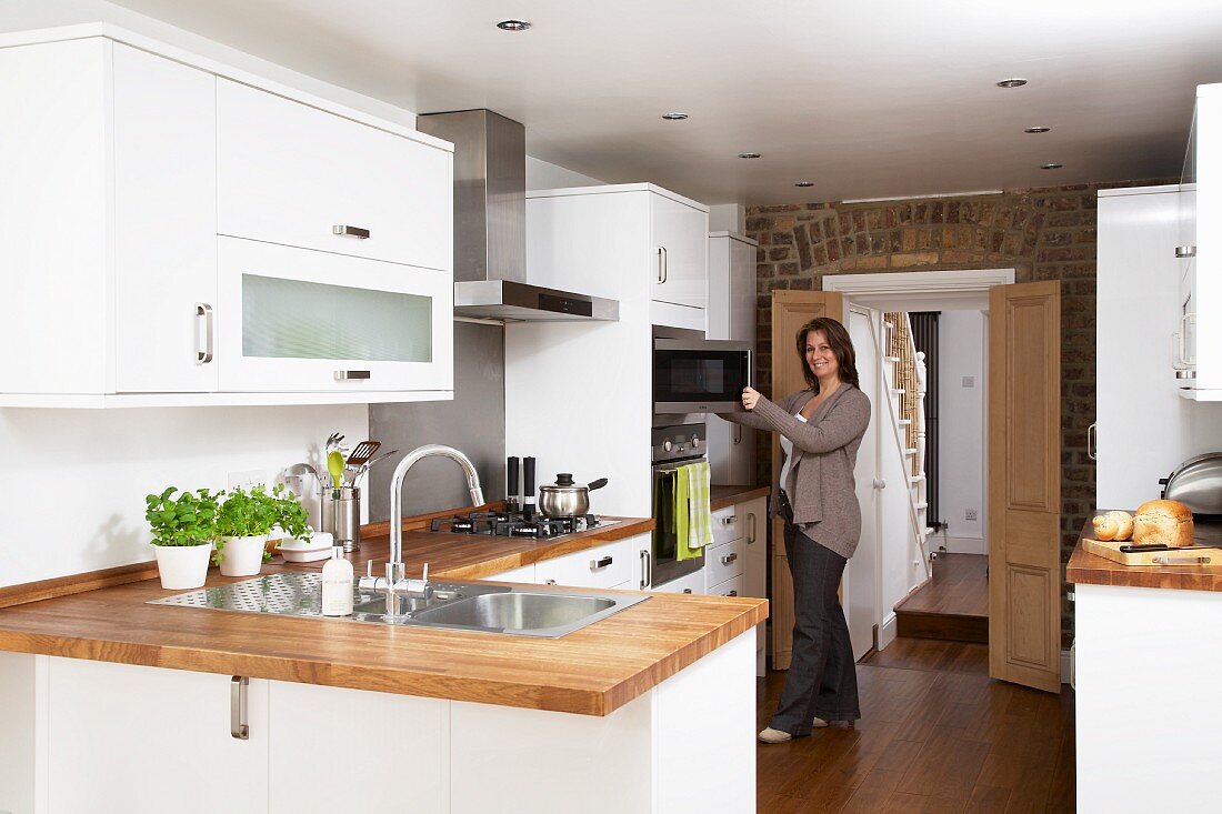 Moderne, weiße Einbauküche mit Arbeitsplatte aus Holz; Frau beim Öffnen der Mikrowelle