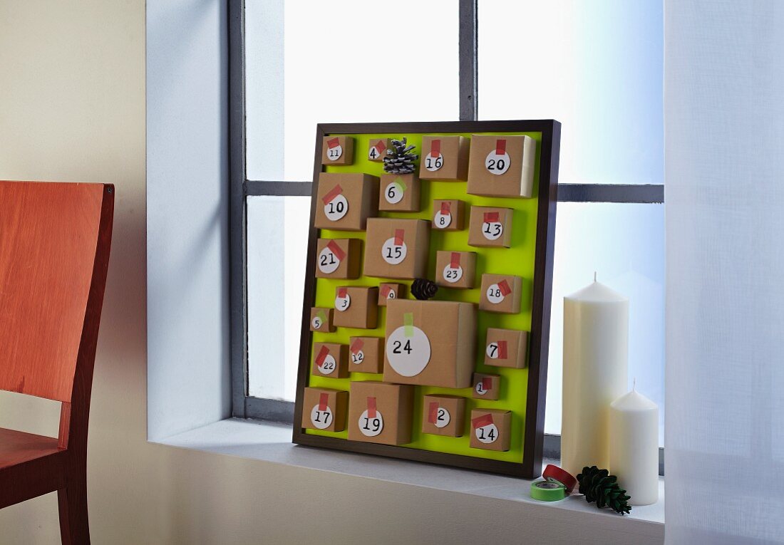 Selbstgebastelter Adventskalender im Bilderrahmen mit kleinen Päckchen auf grüner Rückwand und Kerzen auf Fensterbank