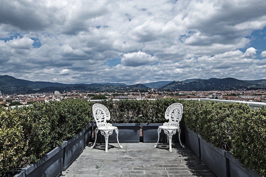 Zwei weiße Vintage Metallstühle im Rokoko Stil in den Ecken der bepflanzten Dachterrasse positioniert mit herrlichem Panoramablick
