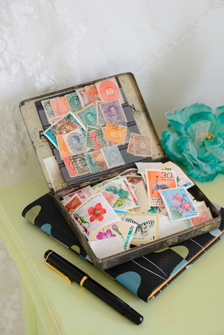 Briefmarken in alter Blechdose auf Notizbuch mit nostalgischem 50er Jahre Design, zwischen Füllhalter und türkiser Seidenblume auf hellgrün lackiertem Tisch