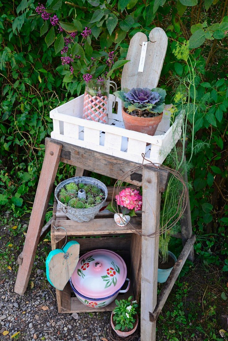 Plants in white crate on rustic side table above nostalgic pots, some planted, on old wooden crate in garden