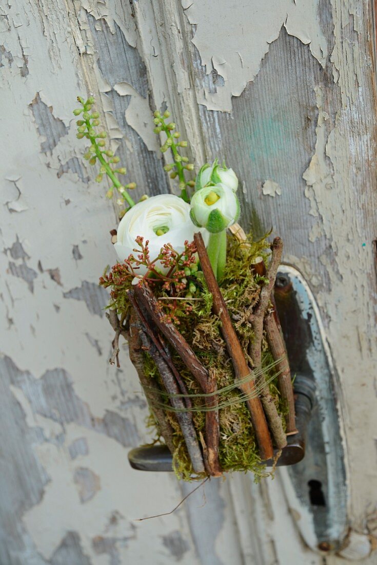 weiße Ranunkelblüten in mit Ästen und Moos umwickelten Marmeladengläschen an einer alten Tür mit abblätternder Farbe