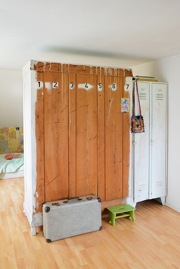 Alter Kleiderschrank und weisser Spind als Raumteiler; Rückseite des Holzschrankes mit Zahlen gestaltet, davor Vintage Koffer und grüner Schemel