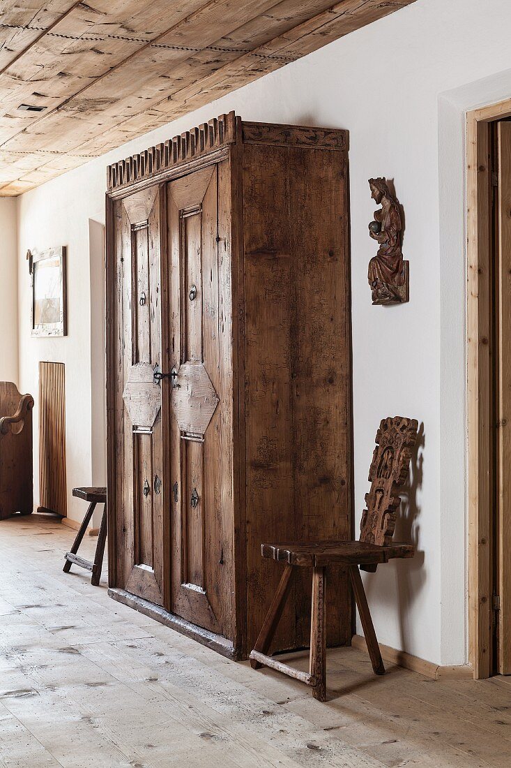 Verzierter, antiker Holzschrank flankiert von zwei Brettstühlen und geschnitzte Heiligenfigur an der Wand