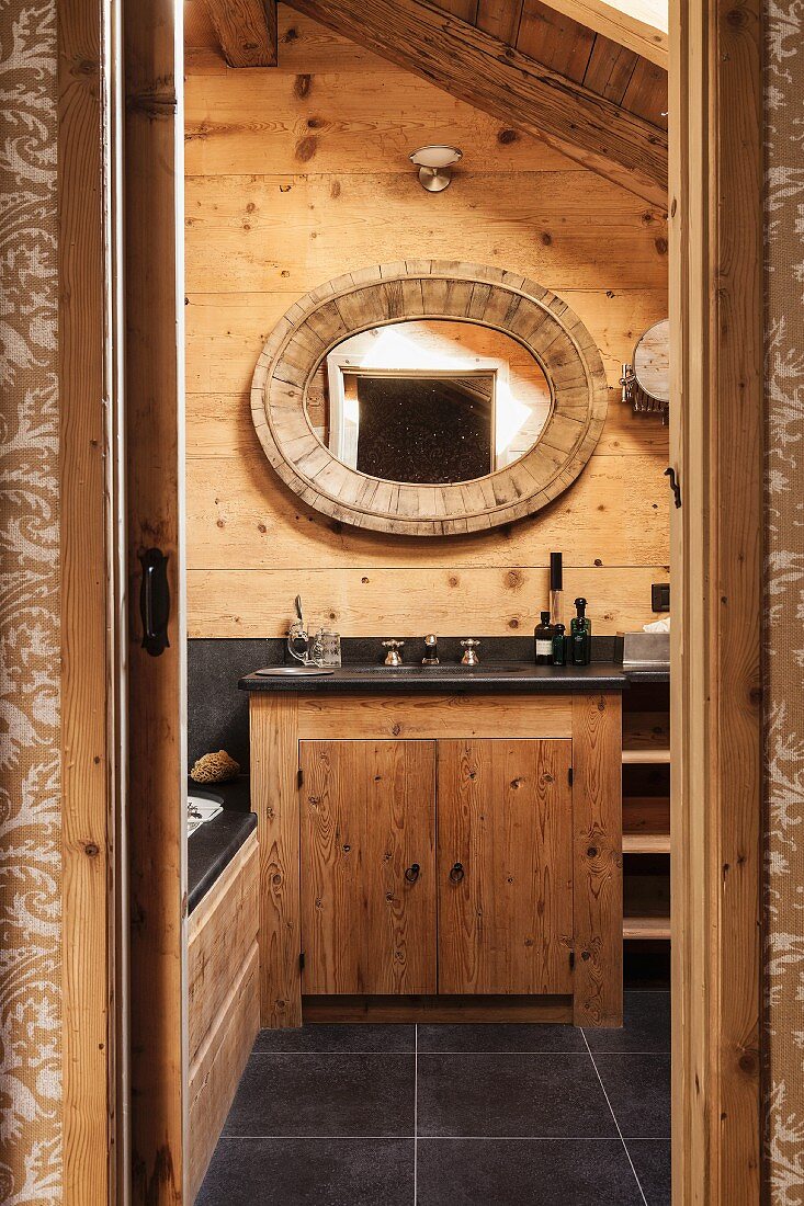 Mit ovalem Holzring gefasster Spiegel an rustikaler Holzwand und integrierter Waschtisch mit schwarzer Ablagefläche