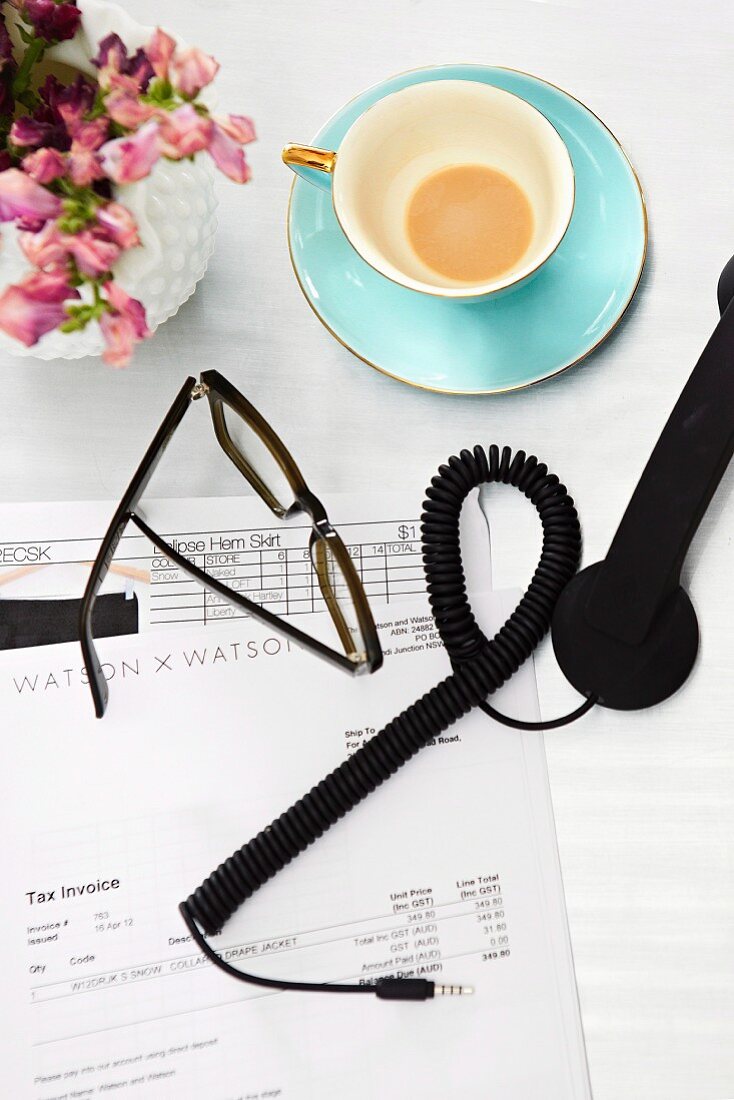 Designerbrille und Telefonhörer auf Firmenbriefpapier, daneben goldrand-Tasse