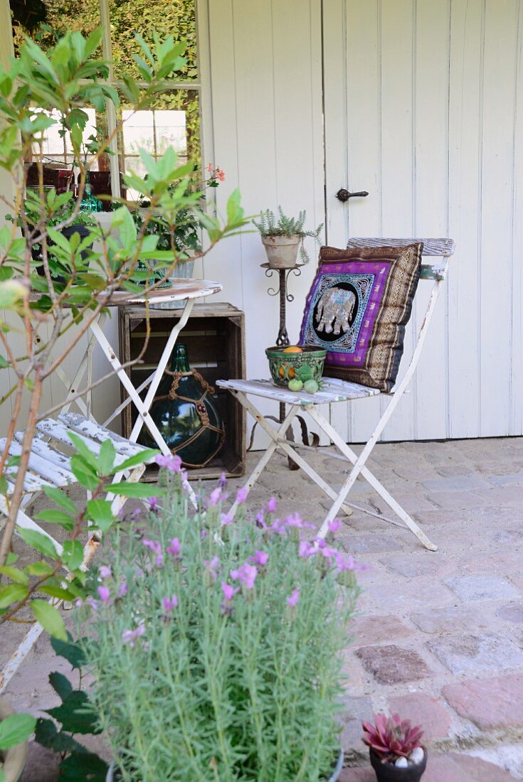 Gepflastertes Terassenplätzchen vor weißem Gartenhaus mit Lorbeer und Lavendel, im Hintergrund alte Klappstühle mit orientalischem Kissen um Tisch