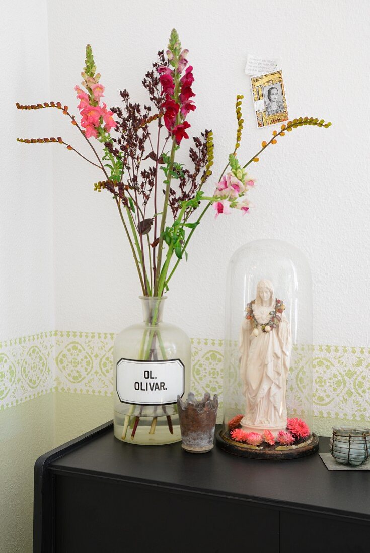 Sommerblumen in Rottönen in Vintage Apothekerflasche, daneben Madonnenfigur dekoriert mit pinkfarbenen Blüten unter Glashaube auf schwarzem Sideboard