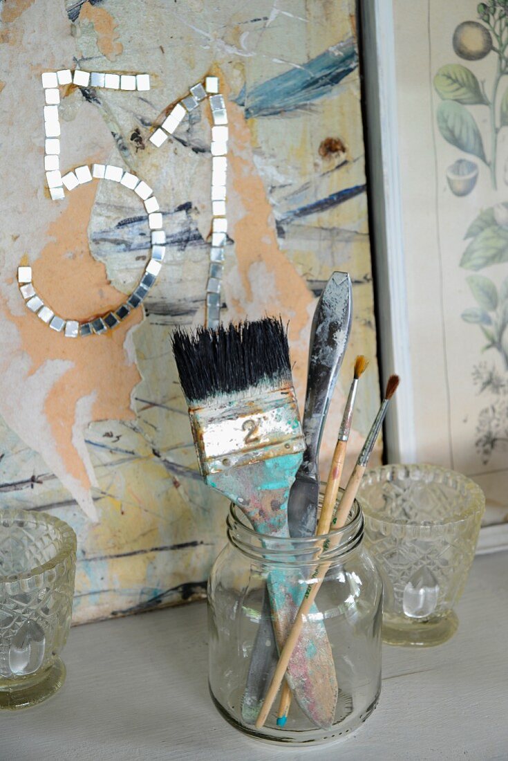Verschiedene Pinsel in Einmachglas zwischen Windlichtern, vor Paneel mit Zahl aus beklebten Spiegel-Mosaikplättchen