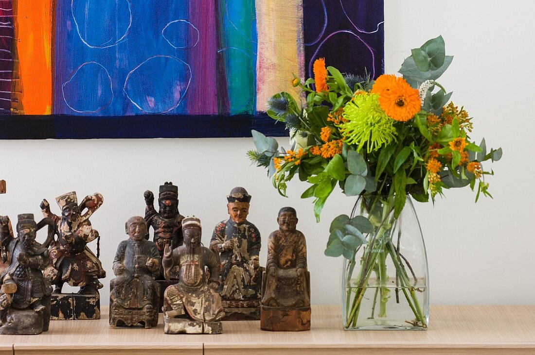 Sammlung von geschnitzten Holzfiguren neben sommerlichen Blumenstrauss vor teilweise sichtbarem Bild an weisser Wand