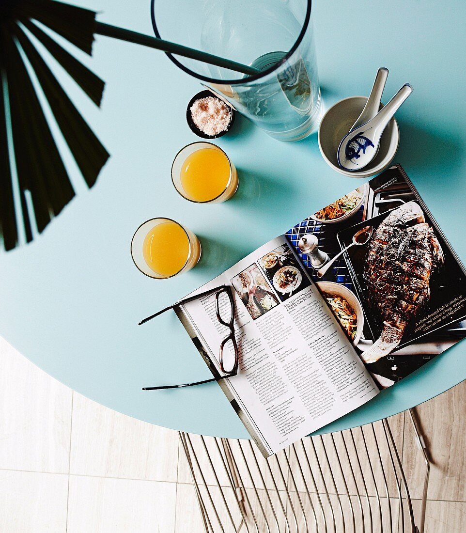 Open cookbook, glasses of juice and palm frond on ice-blue tabletop
