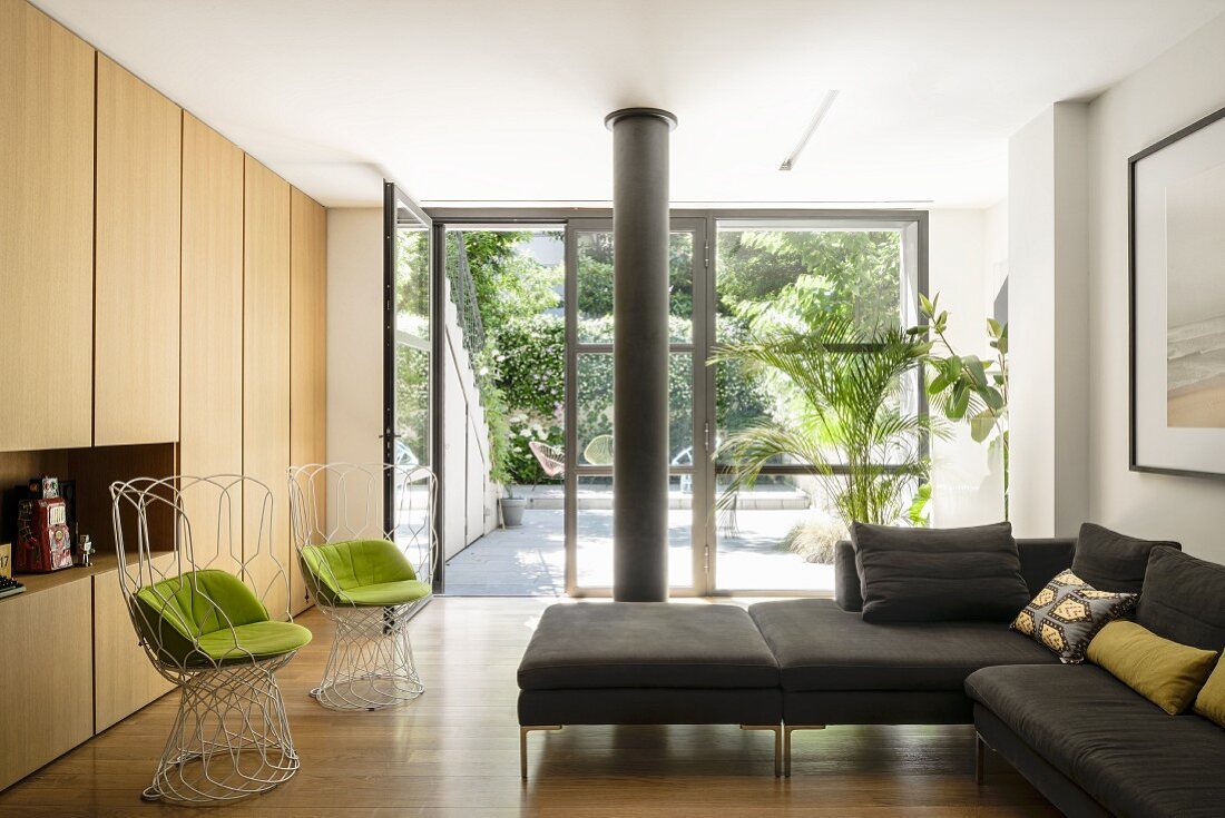 Minimalistisches Wohnzimmer mit modernen Drahtstühlen, hellen Holzfronten und großer Glasfront mit Blick in den begrünten Innenhof
