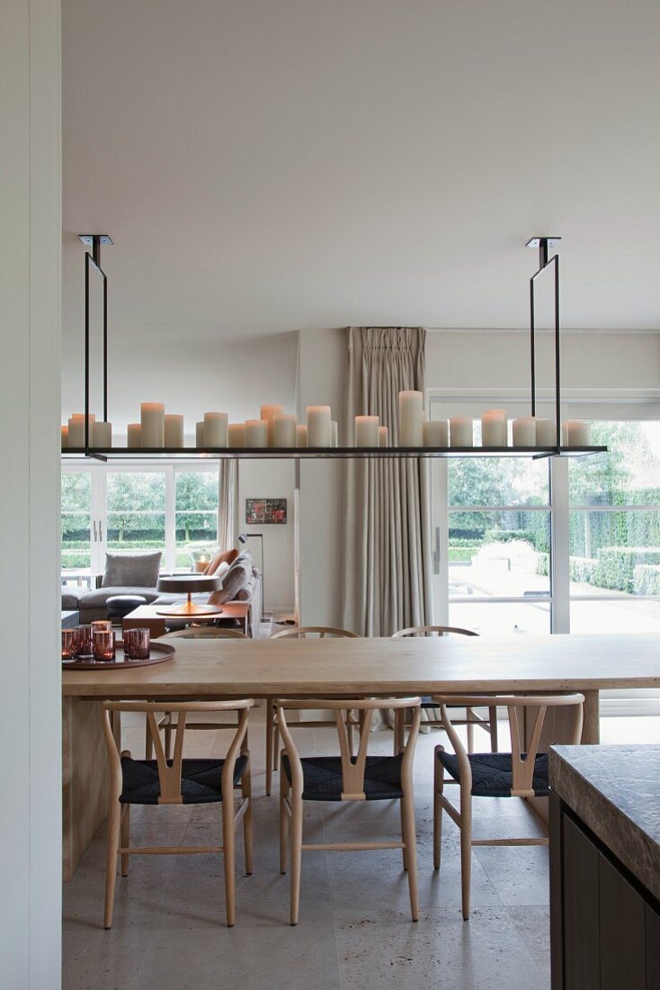 Classic wooden chairs and pale wooden table below collection of white candles on shelf suspended from ceiling; view into garden though French windows in background