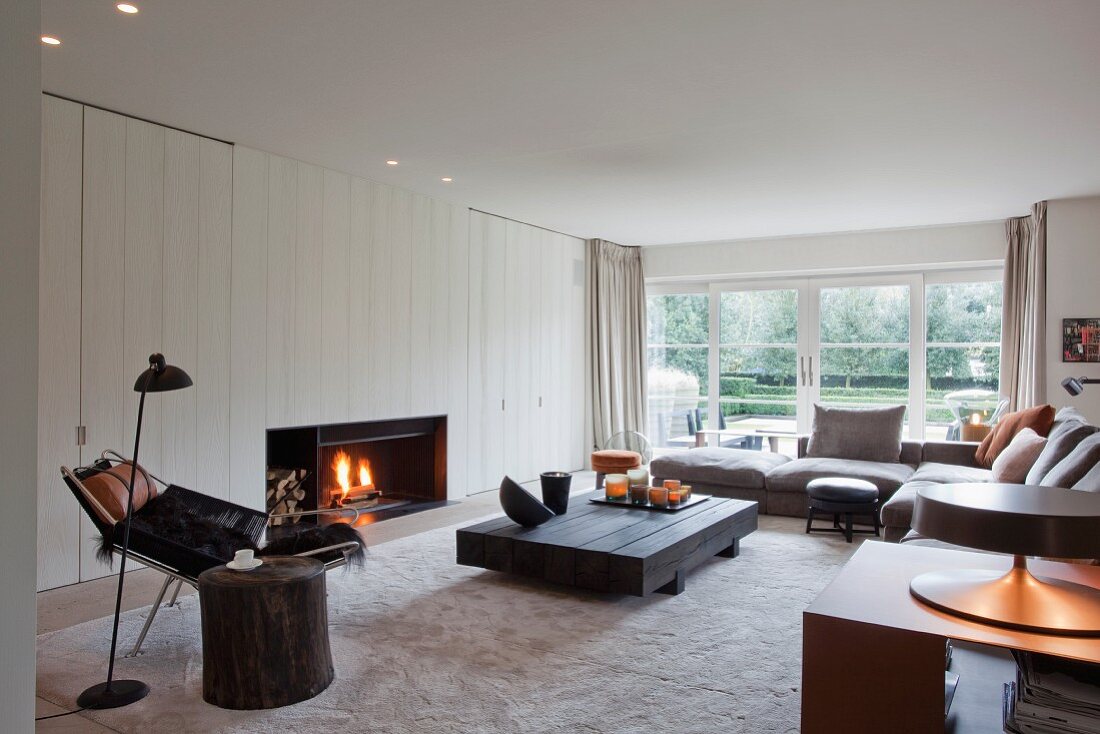 Moderner Wohnraum mit skandinavischem Flair - gemütlicher Loungebereich mit Kaminfeuer, davor Bodentisch aus dunklem Holz und Sofa übereck, seitlich im Vordergrund Leseplatz mit Retro Stehleuchte in Schwarz