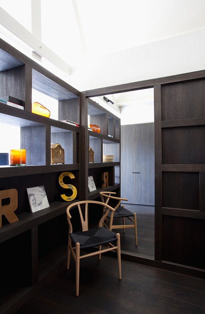Klassikerstuhl aus hellem Holzgestell mit schwarzer Sitzfläche in Zimmerecke, vor schwarzem Holzregal als Raumteiler und gerahmter, raumhoher Spiegel